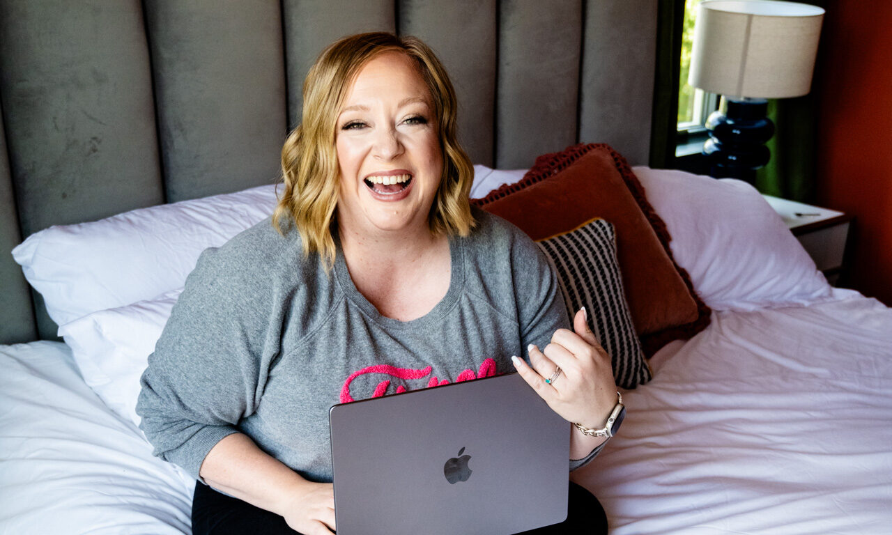 Blonde woman sitting on bed with laptop in hand, looking at camera laughing