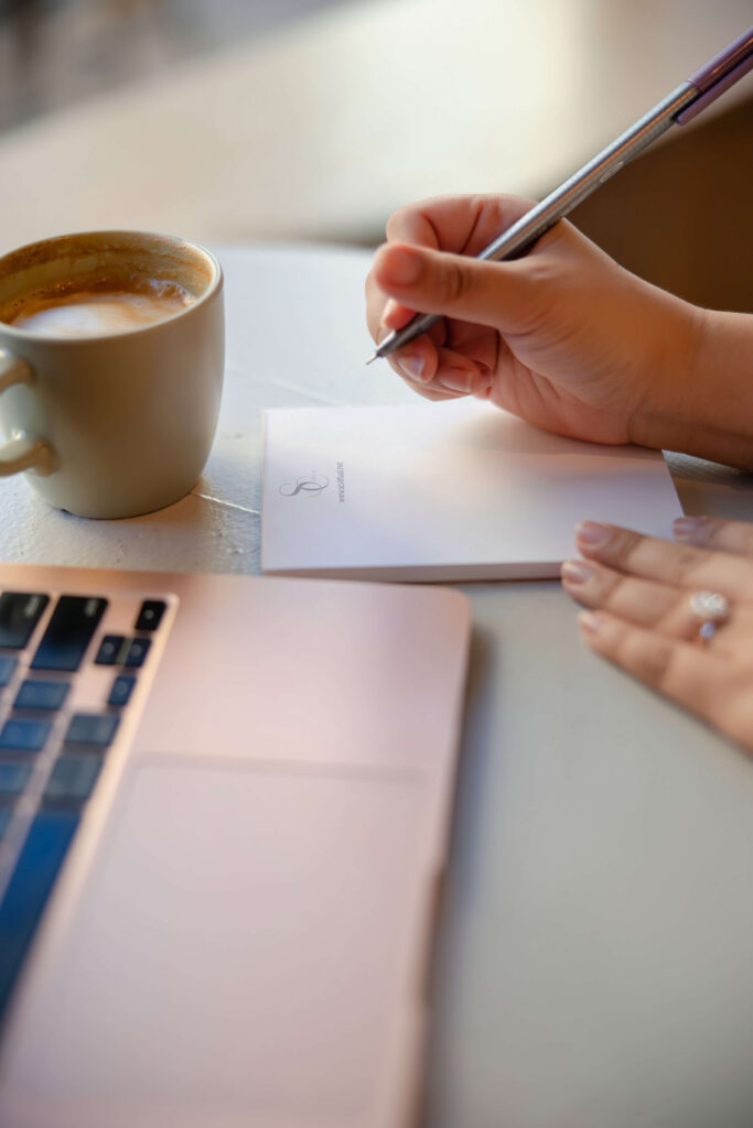 woman's hand writing notes on branded notepad beside open laptop with a cappuccino