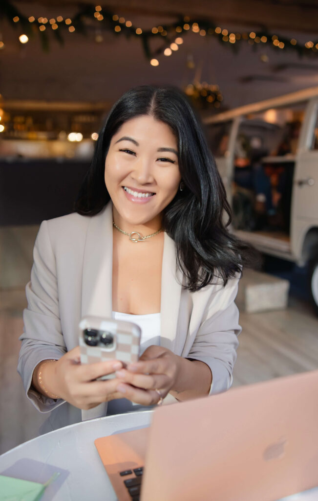 Seattle virtual assistant business woman in beige coat sits smiling while holding her phone looking at camera through window of Pacha Collective coffee shop with a VW van in background