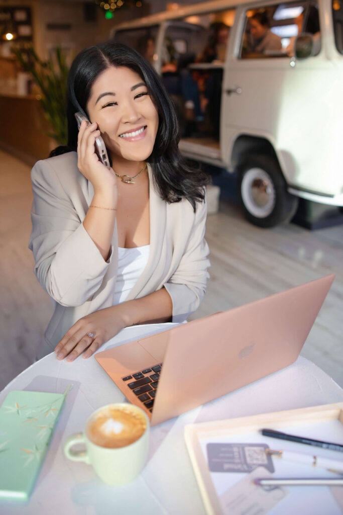 Seattle virtual assistant business woman in beige coat sits at open laptop, smiling while looking at camera through window of Pacha Collective coffee shop with a VW van in background